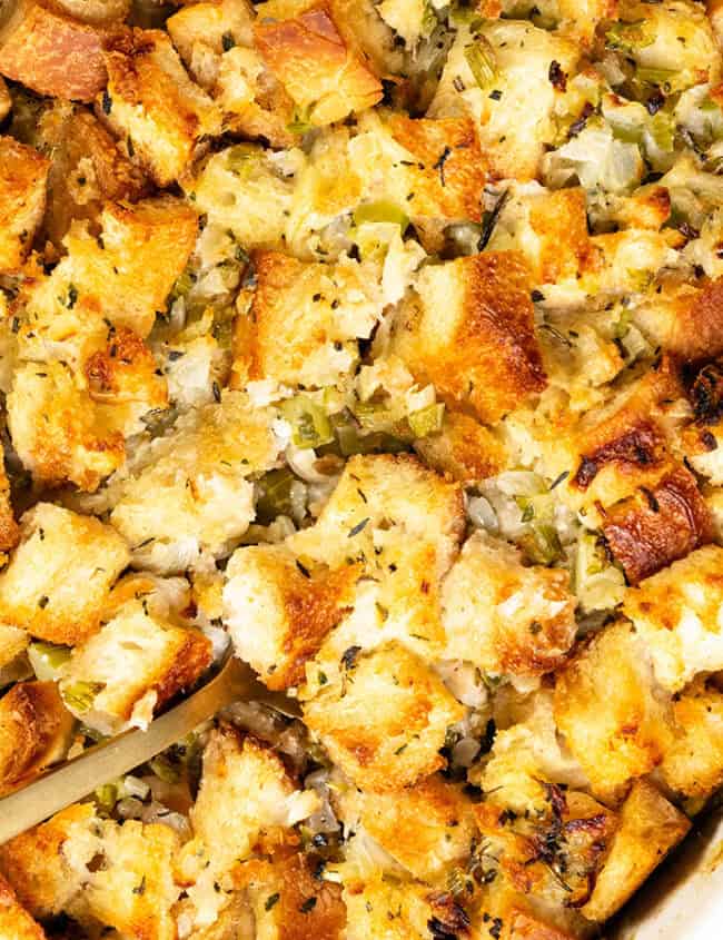 A close-up of a Thanksgiving casserole shows golden-brown bread stuffing, mixed with herbs, onions, and celery. A serving spoon rests in the dish, with fresh sage leaves visible in the corner. A striped cloth is nearby.