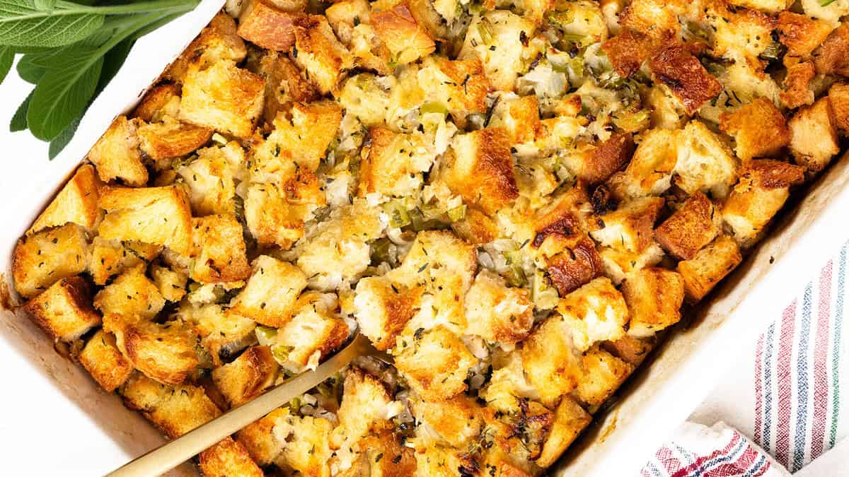 A close-up of a Thanksgiving casserole shows golden-brown bread stuffing, mixed with herbs, onions, and celery. A serving spoon rests in the dish, with fresh sage leaves visible in the corner. A striped cloth is nearby.