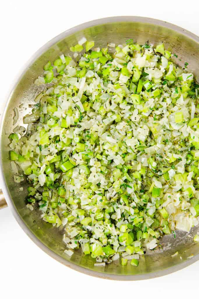 A stainless steel pan contains finely chopped vegetables, including celery, green peppers, and onions, lightly sautéed and evenly distributed for the perfect make-ahead herb stuffing. The mixture has a glistening sheen, suggesting it has been cooked in oil or butter.