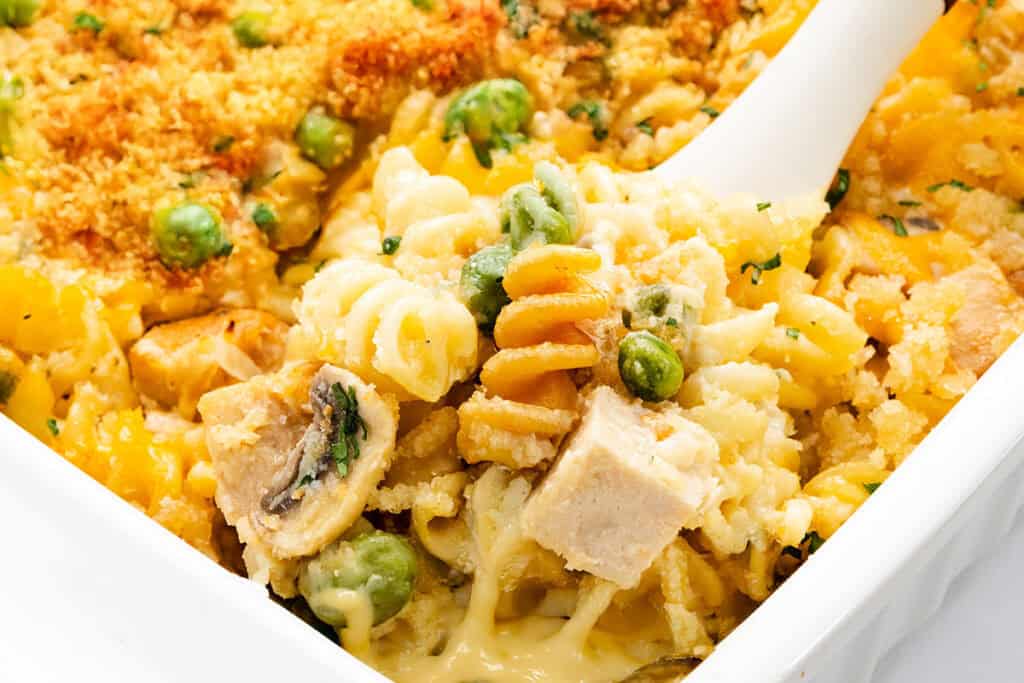 Close-up of a creamy turkey casserole with spiral pasta, chunks of leftover turkey, mushrooms, green peas, and a golden breadcrumb topping. A white serving spoon is scooping a portion from the baking dish.