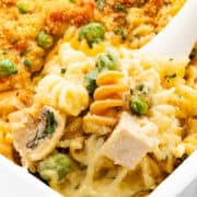 Close-up of a creamy turkey casserole with spiral pasta, chunks of leftover turkey, mushrooms, green peas, and a golden breadcrumb topping. A white serving spoon is scooping a portion from the baking dish.