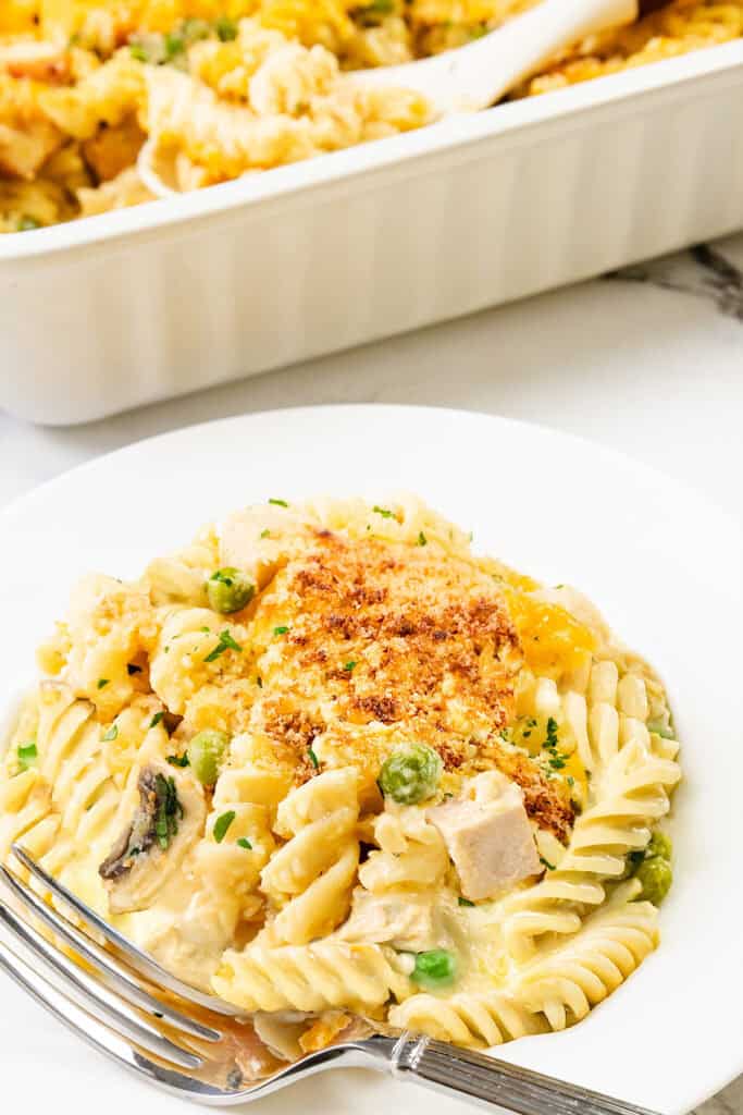 A plate of creamy leftover turkey casserole with rotini pasta, peas, and mushroom slices is topped with golden breadcrumbs and parsley. A serving dish with more casserole is in the background.