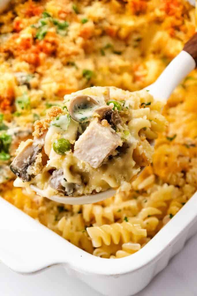 A close-up of a spoonful of creamy pasta bake with mushrooms and leftover turkey, topped with a crispy breadcrumb crust. This turkey casserole features textured rotini pasta and is garnished with chopped parsley, all in a white baking dish.