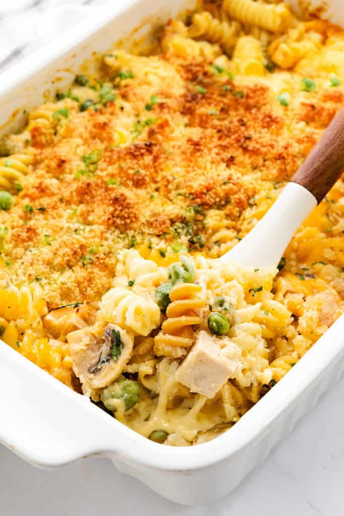 A baked pasta dish with creamy sauce, featuring rotini noodles, peas, and chunks of leftover turkey, topped with golden breadcrumbs, is served in a white casserole dish. A wooden spoon holds a portion of the leftover turkey casserole above the dish.