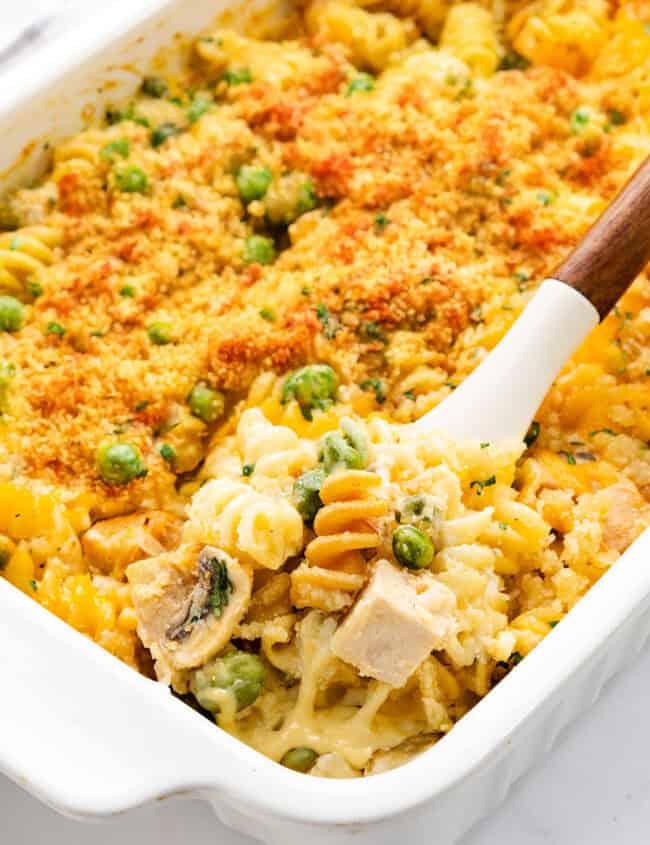 A casserole dish filled with rotini pasta, peas, and chunks of leftover turkey, topped with golden breadcrumbs. A spoon lifts a portion, revealing a creamy cheese sauce underneath.