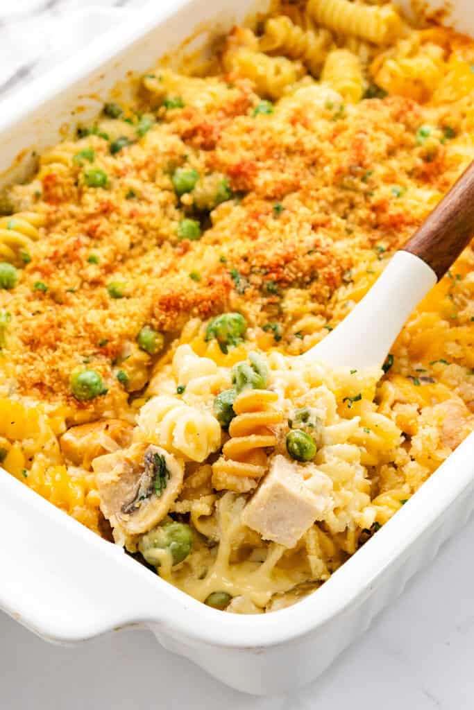 A casserole dish filled with rotini pasta, peas, and chunks of leftover turkey, topped with golden breadcrumbs. A spoon lifts a portion, revealing a creamy cheese sauce underneath.