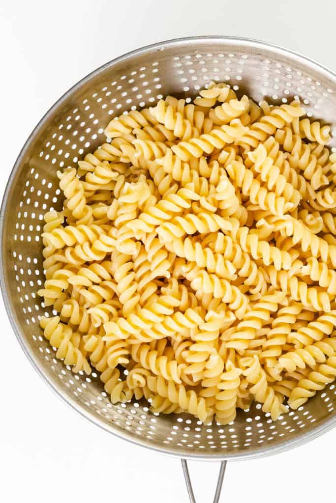 A metal colander filled with cooked rotini pasta, featuring tightly spiraled noodles, is placed on a white background. The pasta, perfect for a turkey casserole recipe, appears to be freshly drained and ready for mixing.