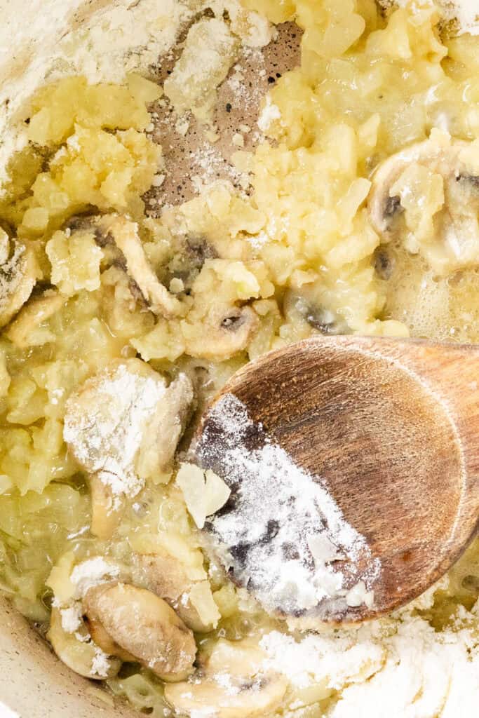 Close-up of a white pot containing sautéed onions and sliced mushrooms being stirred with a wooden spoon. Some flour is sprinkled on top, signaling the start of a roux for a savory turkey casserole, perfect for using up leftovers.