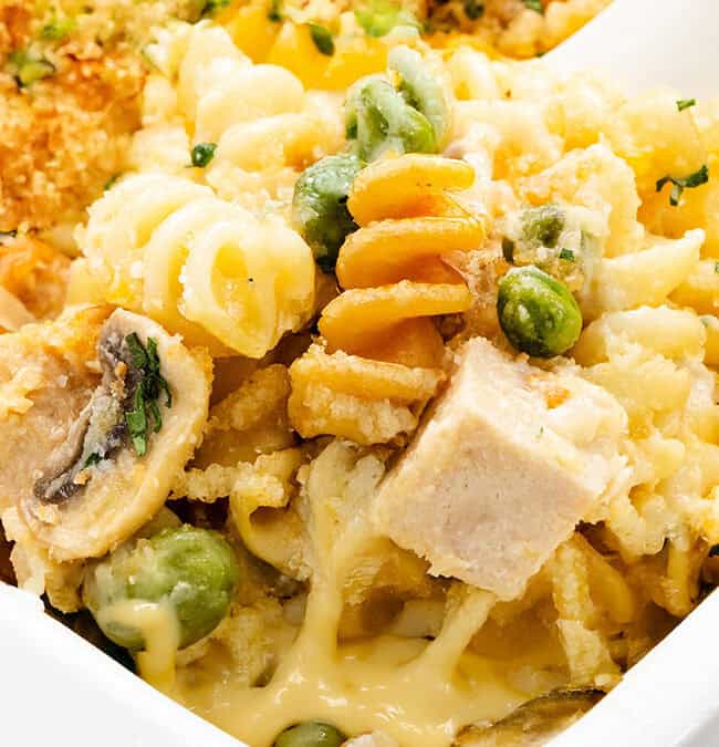 Close-up of a baked pasta dish featuring spiral noodles, cubes of chicken, peas, mushrooms, and a creamy cheese sauce. The top is garnished with breadcrumbs and herbs, creating a golden, crispy texture.