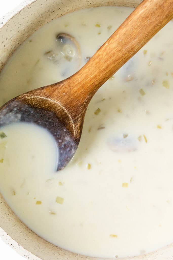 A wooden spoon stirs a creamy white soup with diced vegetables, turkey chunks, and mushroom slices in a pot, transforming leftovers into a delightful casserole-inspired dish.