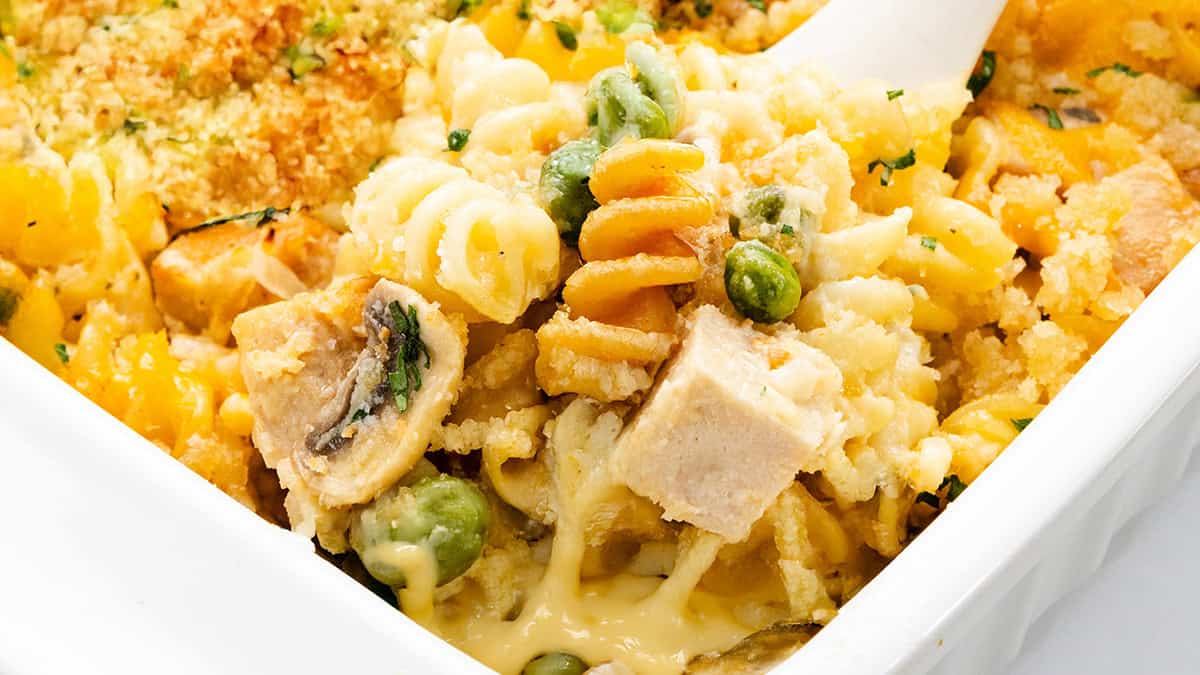 Close-up of a baked pasta dish featuring spiral noodles, cubes of chicken, peas, mushrooms, and a creamy cheese sauce. The top is garnished with breadcrumbs and herbs, creating a golden, crispy texture.