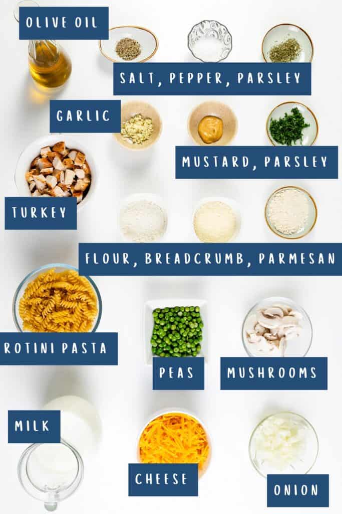 A variety of ingredients arranged on a white surface, each labeled: olive oil, salt, pepper, parsley, garlic, mustard, parsley, turkey for a delicious casserole recipe or to use up leftover turkey. Plus flour, breadcrumb, parmesan, rotini pasta perfect for making a hearty Turkey Casserole with peas and mushrooms.