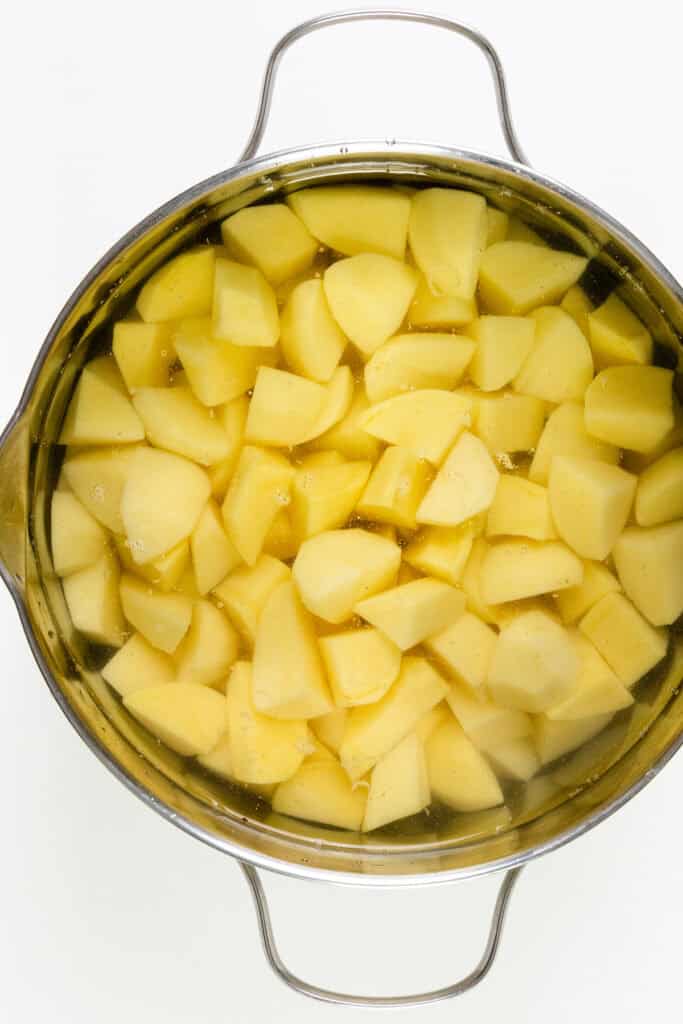 A pot filled with peeled and chopped potatoes, perfect for make-ahead mashed potatoes, is submerged in water. The pot features handles on either side and rests elegantly on a pristine white surface.