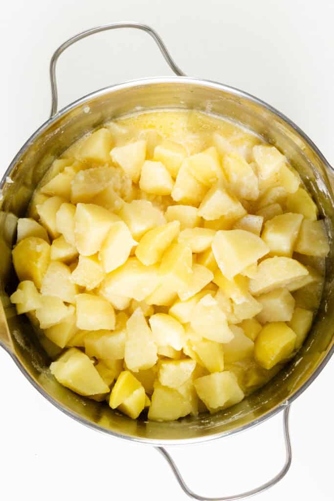 A pot filled with boiled, peeled, and chopped potatoes. These make-ahead mashed potatoes are lightly covered in a buttery or creamy mixture, giving them a slightly glossy appearance. The pot has two handles on either side.