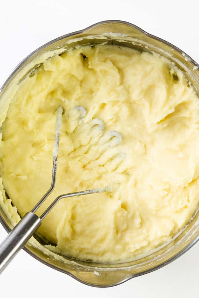 A metal pot filled with creamy, make-ahead mashed potatoes. A metal potato masher is partially visible, pressed into the smooth, fluffy texture of the potatoes, indicating they are being mashed to a soft consistency.