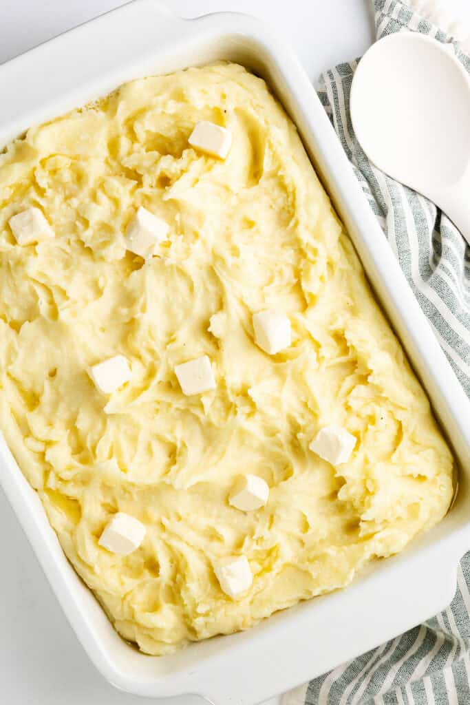 A make-ahead dish of creamy mashed potatoes topped with small cubes of butter sits in a white rectangular baking dish. A striped cloth and a white spoon are elegantly placed beside it, showcasing the perfect comfort food recipe.