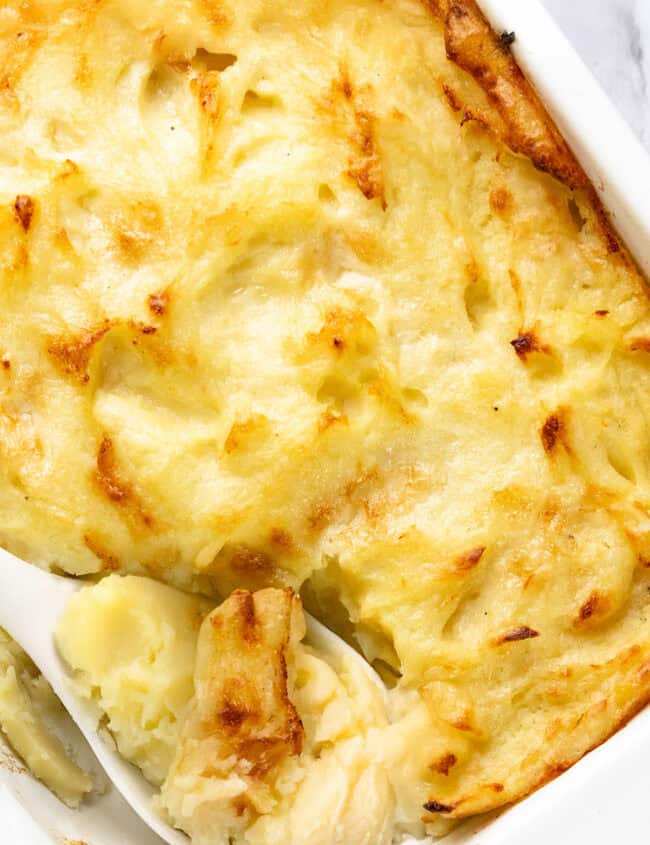 A close-up of a creamy, golden-brown mashed potato casserole in a white baking dish showcases the perfect Make-Ahead Recipe. A white spoon scoops out a portion, revealing the smooth texture of the mashed potatoes beneath the baked top layer.