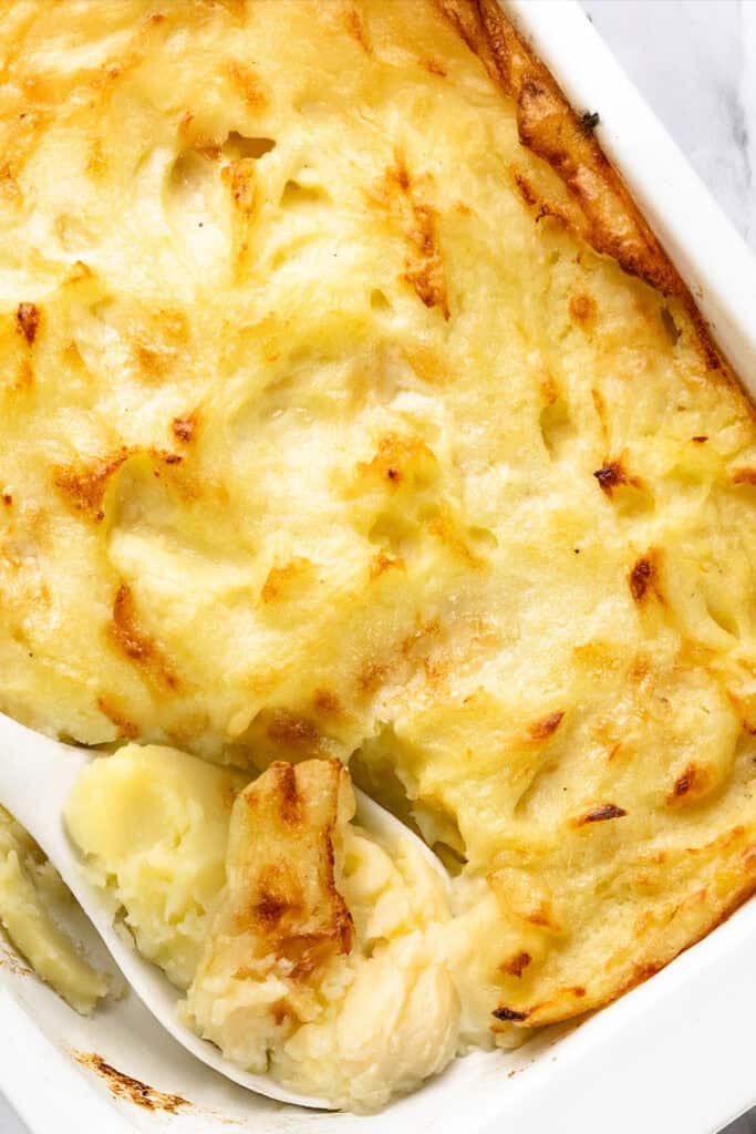 A close-up of a creamy, golden-brown mashed potato casserole in a white baking dish showcases the perfect Make-Ahead Recipe. A white spoon scoops out a portion, revealing the smooth texture of the mashed potatoes beneath the baked top layer.