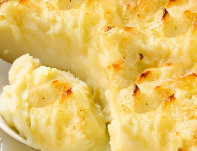 A close-up of a creamy, baked dish of make-ahead mashed potatoes with a golden brown top. A serving spoon is scooping out a portion, revealing the smooth texture beneath the crispy surface.