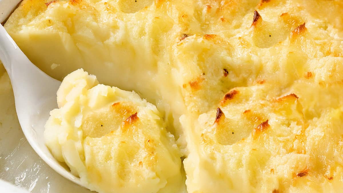 A close-up of a creamy, baked dish of make-ahead mashed potatoes with a golden brown top. A serving spoon is scooping out a portion, revealing the smooth texture beneath the crispy surface.