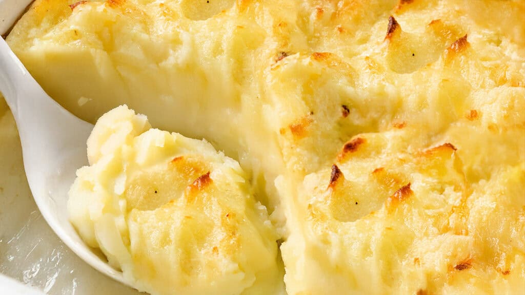 A casserole dish filled with creamy mashed potatoes topped with golden-brown, crispy edges. A serving spoon scoops out a portion, revealing the smooth texture underneath the lightly browned top.