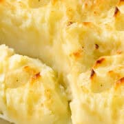 A casserole dish filled with creamy mashed potatoes topped with golden-brown, crispy edges. A serving spoon scoops out a portion, revealing the smooth texture underneath the lightly browned top.