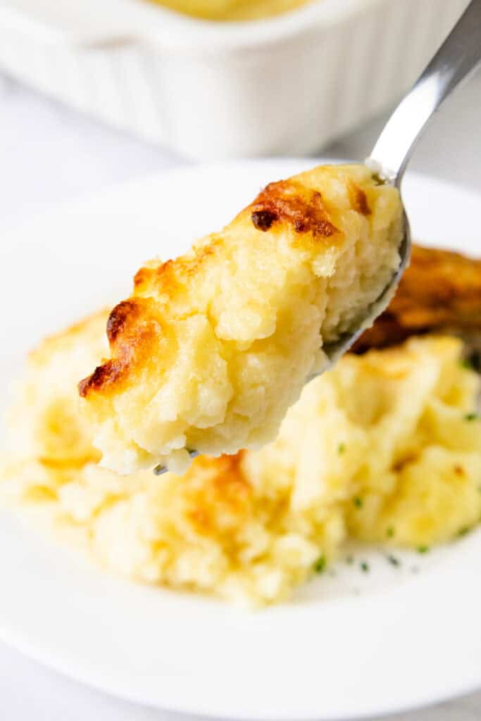 A spoonful of cheesy mashed potatoes, golden brown on top, is held above a plate with more creamy mashed goodness in the background, perfect for those make-ahead meal preparations.