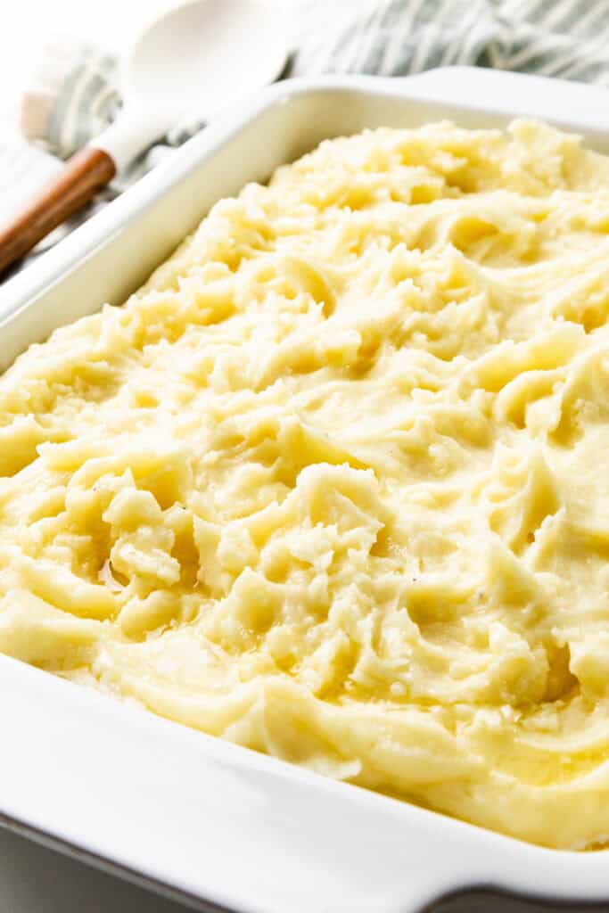 A close-up of creamy, make-ahead mashed potatoes in a white ceramic baking dish. The texture is smooth and fluffy, with a light golden color. In the background, there's a white spoon with a wooden handle resting on a striped cloth.