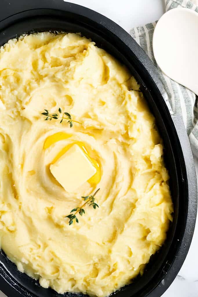 A dish of creamy, make-ahead mashed potatoes topped with a pat of butter and garnished with fresh thyme, served in a black oval dish. A white spoon and a striped cloth are partially visible in the background.