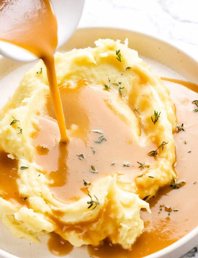 A serving of creamy mashed potatoes topped with Make-Ahead Turkey Gravy being poured from a white bowl. Garnished with sprigs of fresh thyme, the plate is set on a light marble surface.