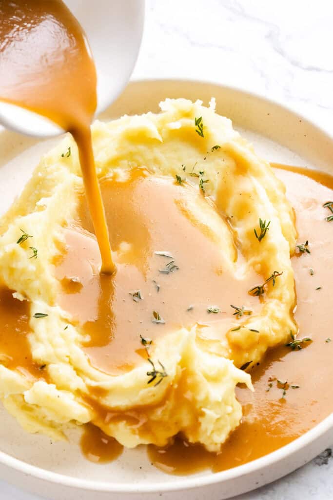 A serving of creamy mashed potatoes topped with Make-Ahead Turkey Gravy being poured from a white bowl. Garnished with sprigs of fresh thyme, the plate is set on a light marble surface.