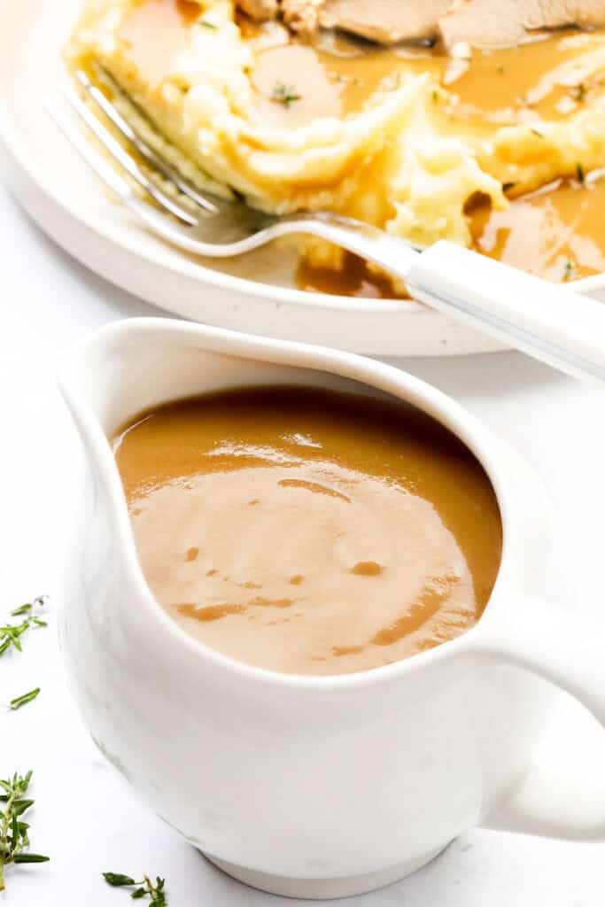 A white gravy boat filled with creamy turkey gravy sits in the foreground. In the background, a plate with mashed potatoes, topped with this make-ahead delight, and a fork can be seen. Small sprigs of herbs are scattered nearby.