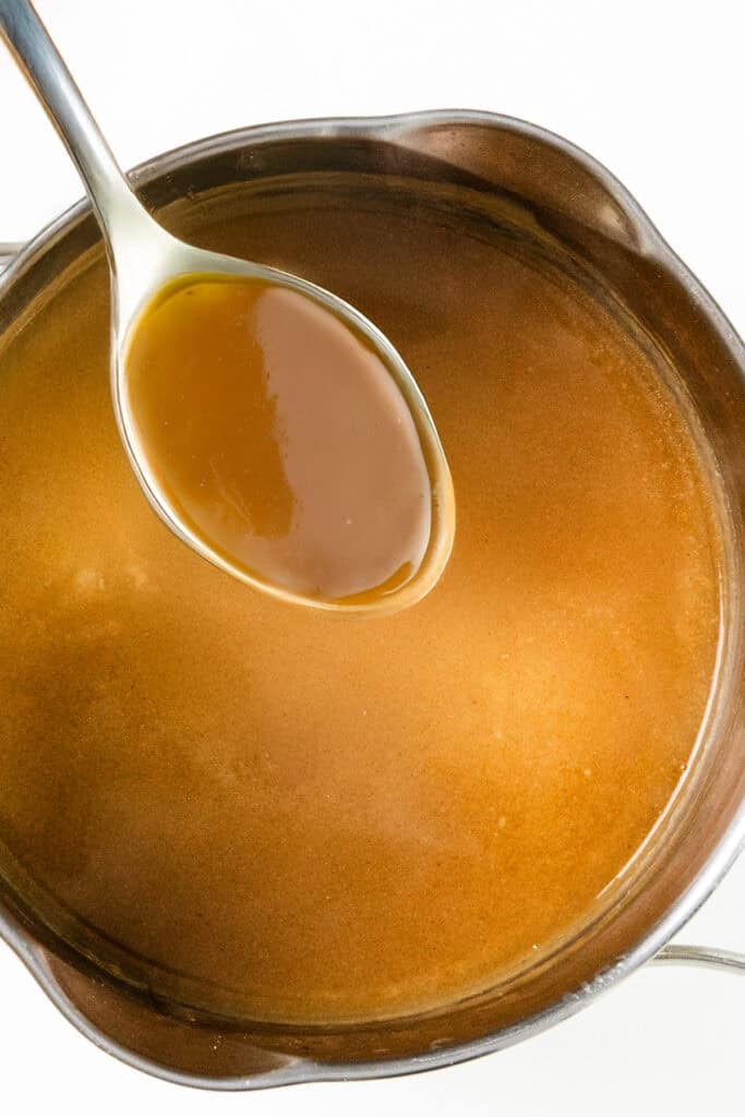 A stainless steel pot filled with smooth, golden brown gravy awaits. A spoon hovers above, showcasing its glossy texture and rich consistency—a perfect component for make-ahead turkey gravy recipes. The clean white background highlights the enticing allure of this thick sauce.