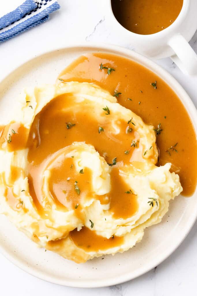 A plate of mashed potatoes topped with savory turkey gravy and garnished with fresh herbs. A gravy boat filled with additional make-ahead gravy is visible in the background. A blue cloth napkin is partially visible on the side.