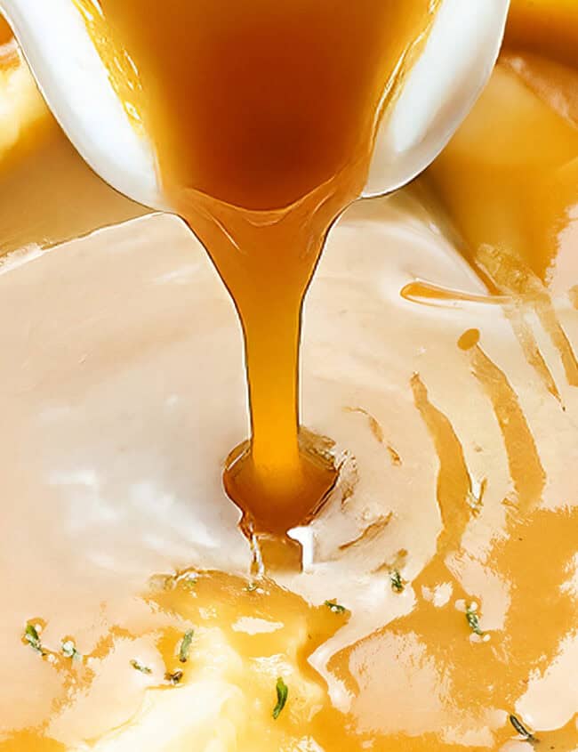 A serving spoon pours golden-brown gravy onto creamy mashed potatoes, a staple in Thanksgiving casseroles, garnished with fresh herbs. The rich sauce forms a small pool on top, highlighting the smooth texture of this festive dish.