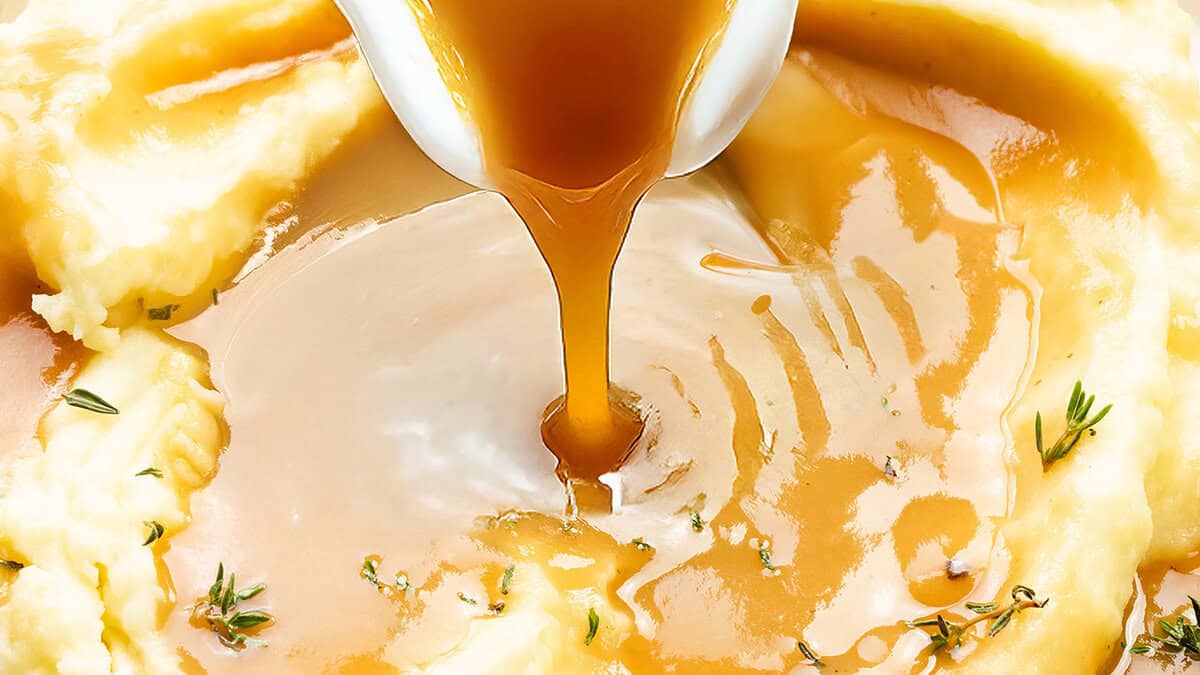 A serving spoon pours golden-brown gravy onto creamy mashed potatoes, a staple in Thanksgiving casseroles, garnished with fresh herbs. The rich sauce forms a small pool on top, highlighting the smooth texture of this festive dish.