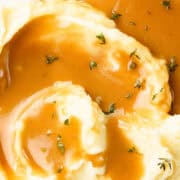 A plate of creamy mashed potatoes topped with rich, make-ahead turkey gravy and garnished with sprigs of fresh thyme. The dish appears smooth and appetizing, inviting a comforting meal experience.