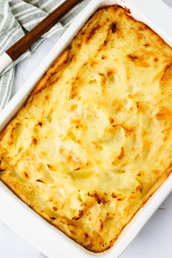 A make-ahead mashed potato casserole in a rectangular white dish, topped with a golden-brown crust. A wooden-handled utensil rests on a folded striped cloth beside the dish.