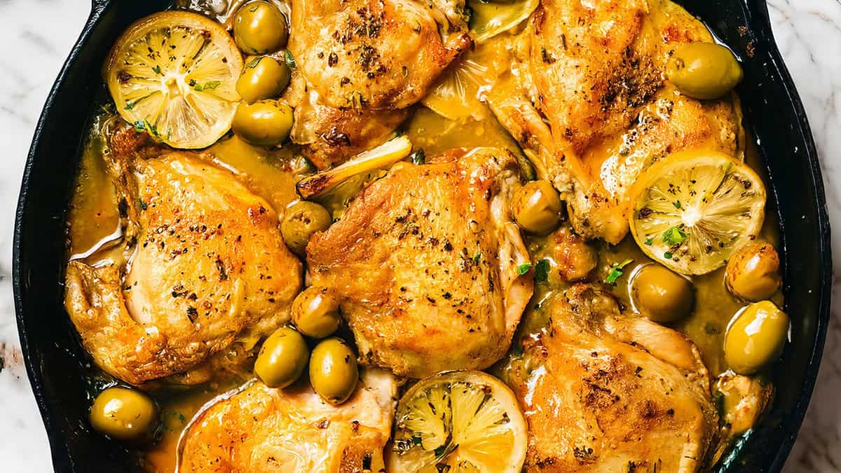 Close-up of a skillet showcasing a tried-and-true recipe with roasted chicken thighs, sliced lemons, and green olives. The dish appears to be seasoned with herbs and spices, hitting the mark with its golden-brown finish and savory, well-cooked appearance.