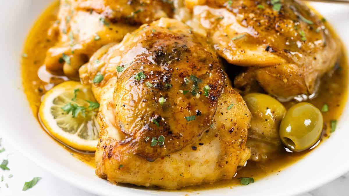 A close-up of seasoned, cooked chicken pieces served in a flavorful sauce with olives and lemon slices, garnished with fresh herbs—true comfort food at its finest.