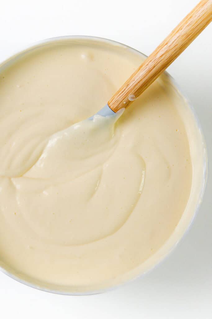 A bowl of creamy off-white batter, hinting at a luscious New York Cheesecake, with a wooden-handled spatula resting on its surface. The mixture is smooth and dense, ready for baking. The white background accentuates the texture and inviting color of the batter.