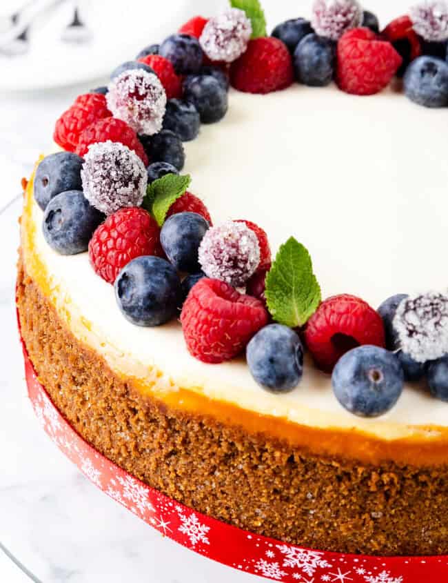 This New York cheesecake is topped with fresh blueberries, raspberries, and sugared cranberries. The crust is golden brown, and the cake is wrapped with a festive red ribbon. The dessert rests on a white surface, with two blurred spoons in the background.