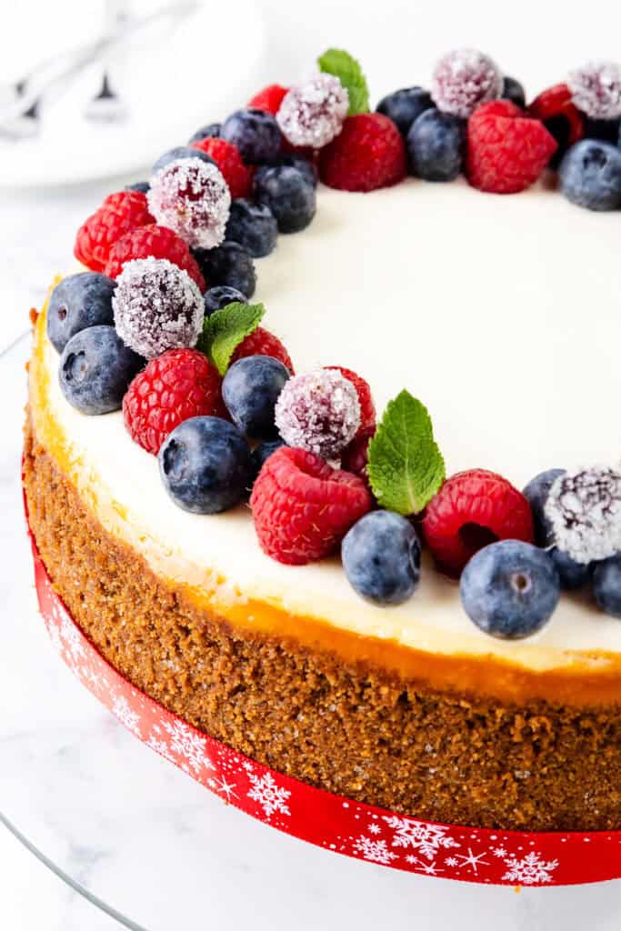 This New York cheesecake is topped with fresh blueberries, raspberries, and sugared cranberries. The crust is golden brown, and the cake is wrapped with a festive red ribbon. The dessert rests on a white surface, with two blurred spoons in the background.