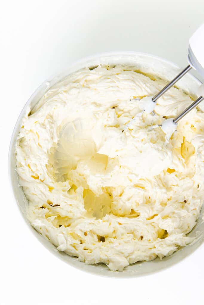 A bowl filled with whipped cream cheese frosting, reminiscent of a classic New York cheesecake, is being mixed with an electric hand mixer. The beaters are partially visible, surrounded by smooth, creamy frosting.