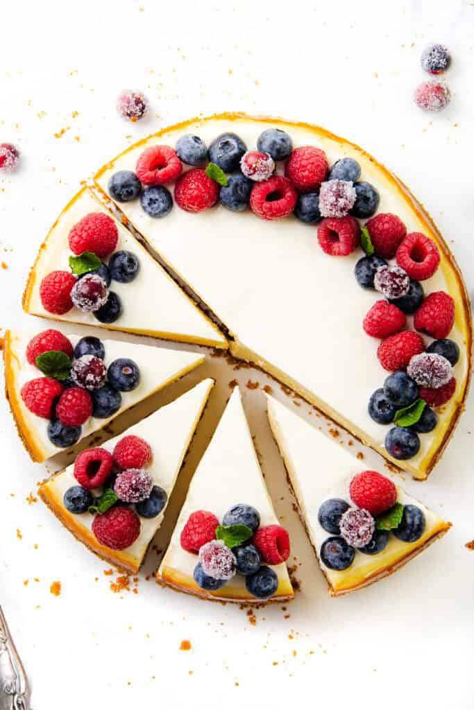 A New York cheesecake topped with fresh raspberries and blueberries, featuring a crumbly crust. Several slices are cut, revealing its creamy texture. The cake is elegantly displayed on a white surface, with a silver serving utensil nearby.