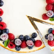 A creamy New York cheesecake topped with fresh blueberries, raspberries, and mint leaves. A slice is being pulled away, revealing the smooth texture inside. It’s beautifully decorated with a golden-brown crust on a marble surface.