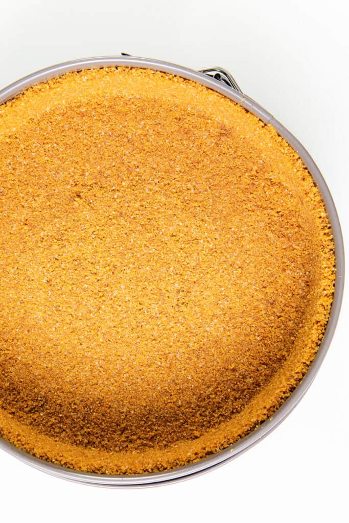 A close-up of a golden-brown, crumbly cheesecake crust in a round baking pan, viewed from above. The New York-style crust appears evenly baked with a textured surface, set against a plain white background.