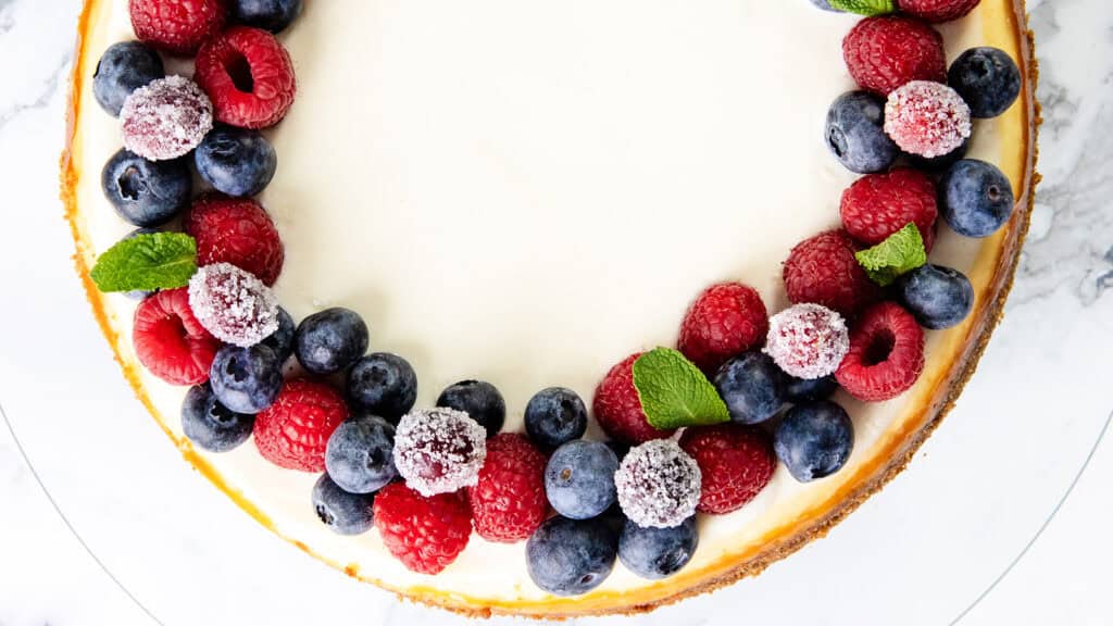 A cheesecake garnished with a circular arrangement of fresh blueberries and raspberries. Some berries are dusted with sugar, and small mint leaves are interspersed among the fruits. The cake sits on a white surface.