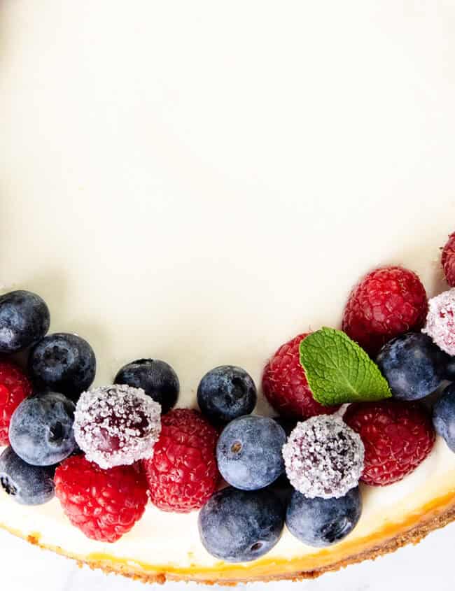 A cheesecake garnished with a circular arrangement of fresh blueberries and raspberries. Some berries are dusted with sugar, and small mint leaves are interspersed among the fruits. The cake sits on a white surface.