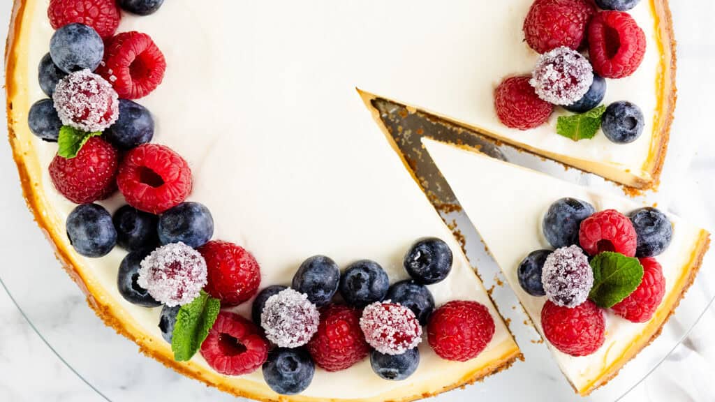 A New York cheesecake topped with fresh raspberries, blueberries, and mint leaves. A slice has been cut from the cake, revealing a creamy interior. The cake is elegantly displayed on a white marble surface.